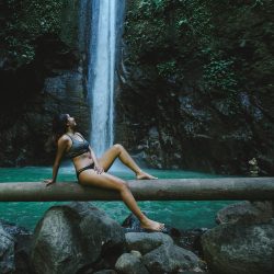 A beautiful woman stands beneath a large, stunning waterfall with emerald green water in a native Amazon rainforest. An incredible landscape.